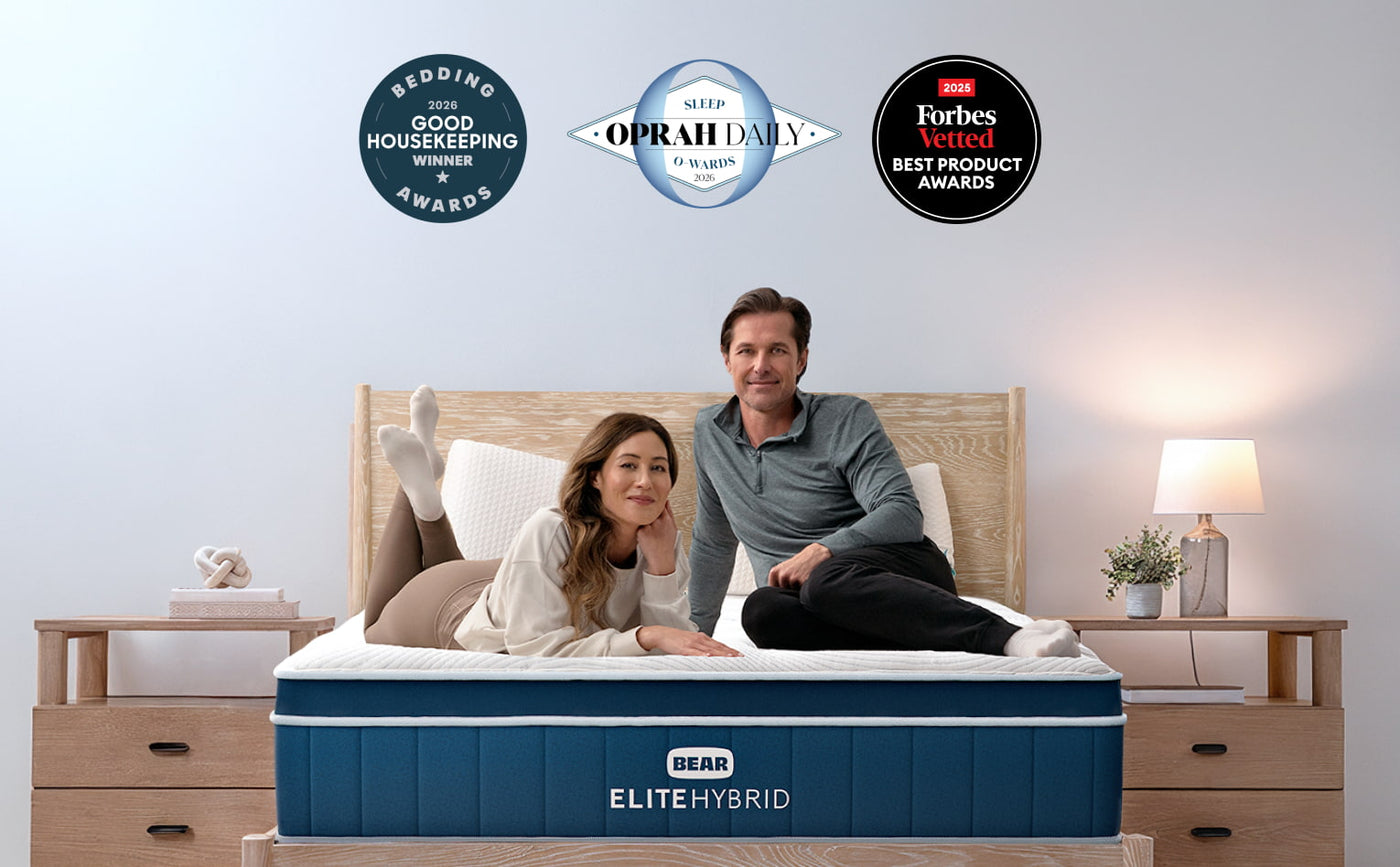 A man and a woman sit on a BEAR ELITE HYBRID mattress in a styled bedroom setting. The mattress features a blue and white design with the BEAR brand logo.