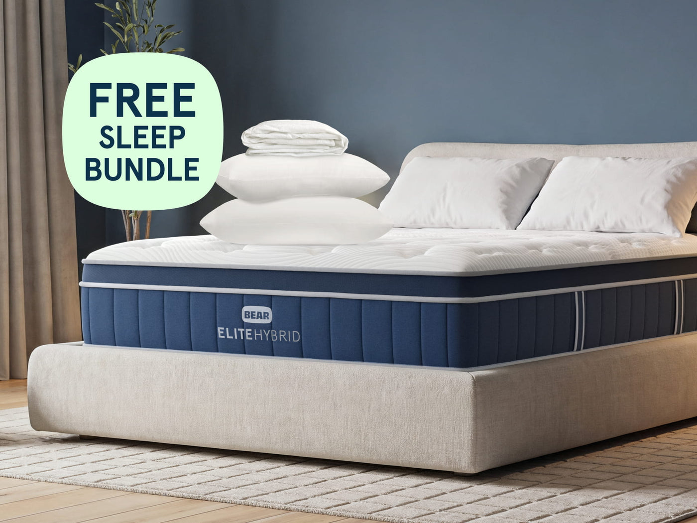 A bed with a navy and white ELITE HYBRID mattress, styled with white pillows and folded sheets on top, in a bedroom setting with a blue wall and a light green 'FREE SLEEP BUNDLE' badge overlay.