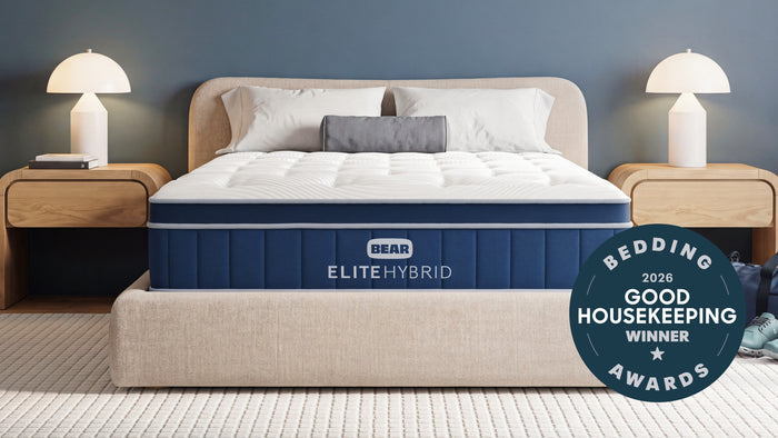 A BEAR ELITE HYBRID mattress shown on a bed in a styled bedroom setting with a blue wall, bedside tables, lamps, and a circular 'Bedding 2026 Good Housekeeping Winner Awards' badge on the right.
